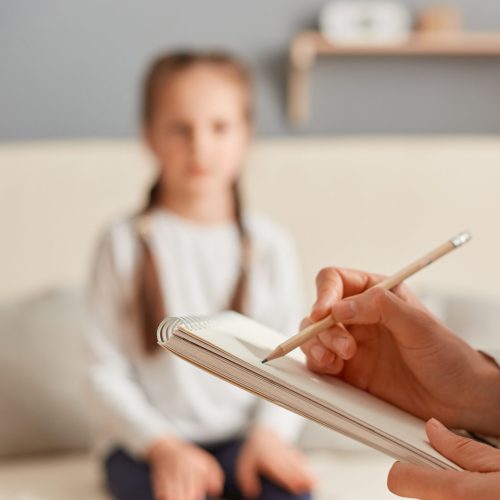 Closeup of doctor psychologist holding paper notebook making notes while talking with small girl at psychological therapy session meeting, blur defocused little kid patient.
